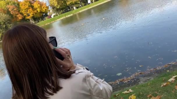 Jonge vrouw met smartphone zit aan de vijver in het najaarspark. — Stockvideo