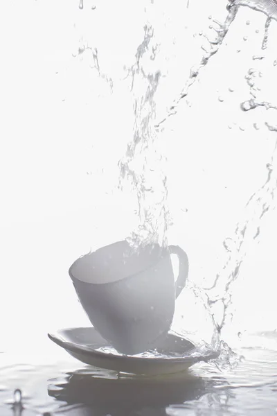 White cup and saucer with splashes of water.Vertical photograph of glass and water on a white background.