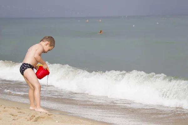 赤ちゃんは 海のそばのバケツから水を注ぐ 少年は 海岸で遊ぶ 子供の夏休みは海岸ではないです — ストック写真