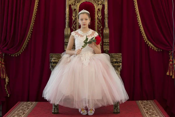 Teenager girl smart in red luxury royal armchair. Girl princess.Queen in a red chair.Luxurious young lady with a scarlet rose in a white dress in an old red armchair