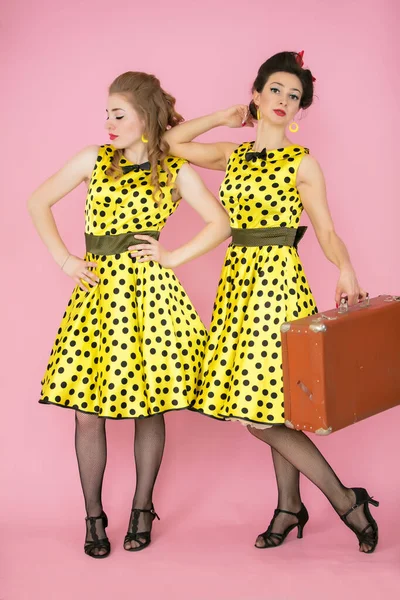 Retro lányok egy bőrönddel. Két nő sárga ruhában, rózsaszín háttérrel.. — Stock Fotó