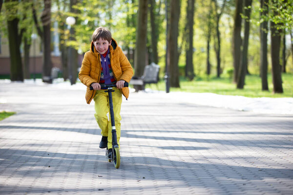 The child rides a scooter in the park. A boy in bright clothes plays a spring day.