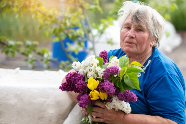 Gray-haired elderly woman with a bouquet of lilacs outdoors. Pensioner in the country. — Stock Photo, Image