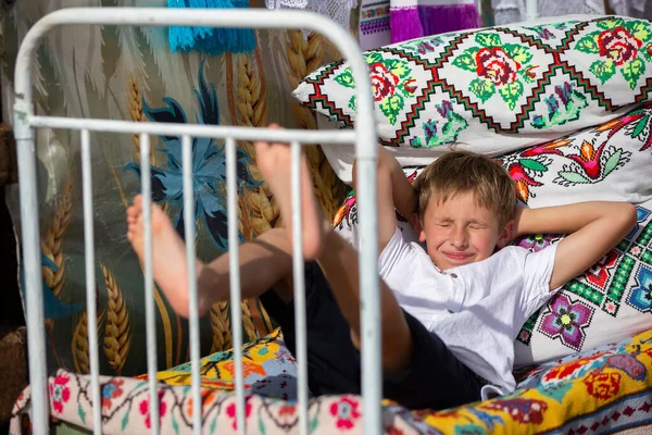 Ребенок русский или украинский на старой этнической кровати с подушками и покрывалами, вышитой славянским узором. Белорусский мальчик. — стоковое фото
