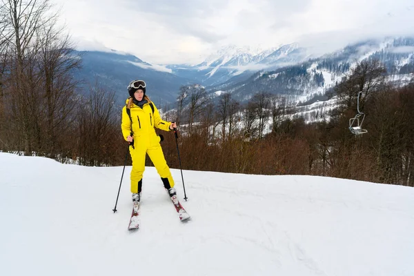 Parlak sarı kayak takımlı bir kadın, karlı dağ zirvelerine karşı poz veriyor. Kış tatili atmosferi ve aktif turizm.