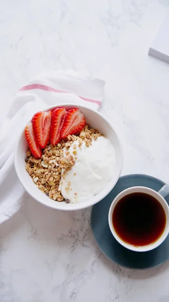 Perfect breakfast of crunchy granola with yoghurt and strawberries in bowl and cup of black coffee on marble table