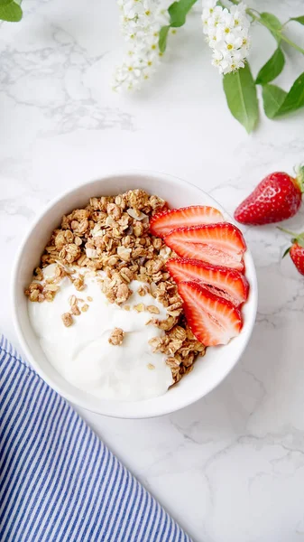 crunchy granola with yoghurt and strawberries in white bowl with bird-cherry tree flowers , close-up