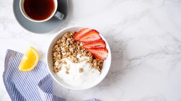Perfect breakfast of crunchy granola with yoghurt and strawberries in bowl and cup of black coffee on marble table