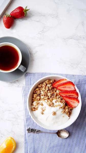 Perfect breakfast of crunchy granola with yoghurt and strawberries in bowl and cup of black coffee on marble table