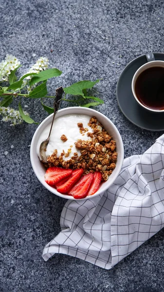 Yoğurtlu gevrek gevrek ve çilek ile fincan sade kahve ve gri arka plan, mükemmel kahvaltı kavramı üzerinde çiçekler ile kase 