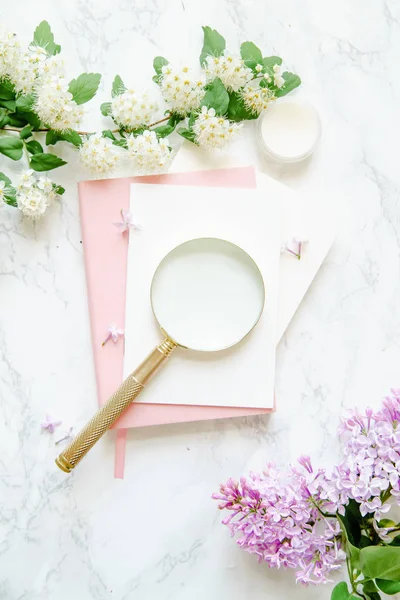 ピンクのノートと大理石の背景 女性ブロガー ワークスペースの概念に黄金の拡大鏡を白とライラックの花クリーム — ストック写真
