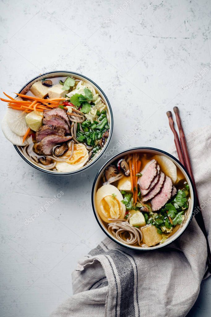 Bowls of ramen with roasted beef, shiitake mushrooms, fried tofu, leek and eggs on concrete background