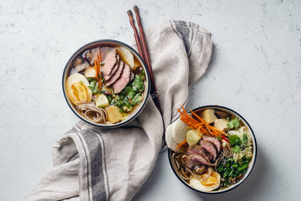 Bowls of ramen with roasted beef, shiitake mushrooms, fried tofu, leek and eggs on concrete background