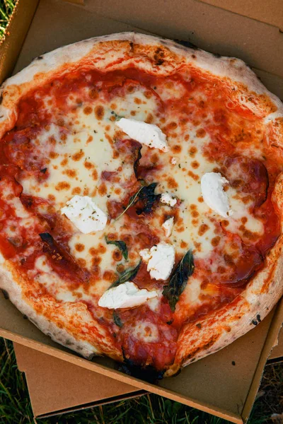 Italian pizza with tomato sauce and fresh burrata in opened cardboard box on grass, Food delivery concept