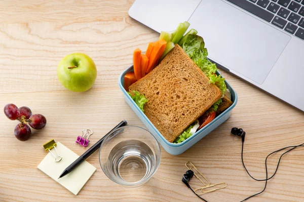 healthy sandwich in plastic container with fruits and office items next to opened laptop on wooden table. Takeaway food for lunch at work
