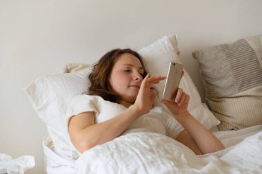 Sabahları yatakta yatarken akıllı telefondan mesajlar okuyan genç bir kadın. 