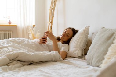 Sabahları yatakta yatarken akıllı telefondan mesajlar okuyan genç bir kadın. 