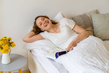 Sabahın erken saatlerinde yatakta oturmuş roman okuyan genç bir kadın.