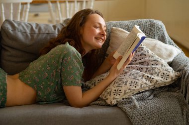 Evdeki kanepede uzanmış kitap okuyan genç bir kadın. 