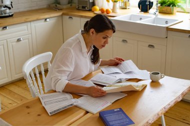 Genç bir kadın mutfakta kitap ve doküman kullanırken masada oturup ders çalışıyor. 