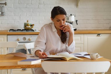 Genç bir kadın mutfakta kitap ve doküman kullanırken masada oturup ders çalışıyor. 