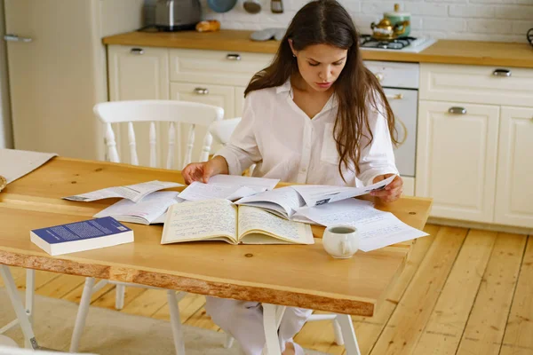 Young woman sitting at table and studying while using notepads with books and documents in kitchen