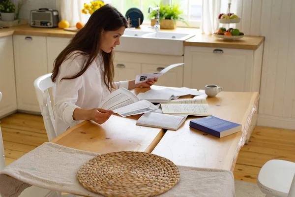 Genç bir kadın mutfakta kitap ve doküman kullanırken masada oturup ders çalışıyor. 