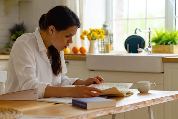 Young woman sitting at table and studying while using notepads with books and documents in kitchen