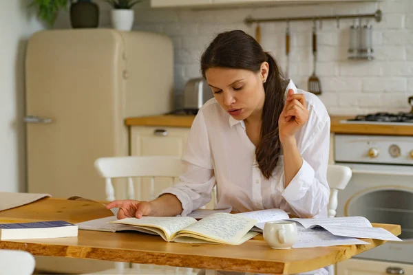 Young woman sitting at table and studying while using notepads with books and documents in kitchen