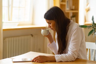 Mutfakta Tablet PC ile masada otururken beyaz fincan kahve içme genç kadın 