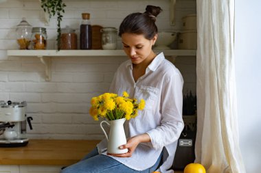 Mutfakta masada otururken parlak sarı çiçek buketi ile vazo tutan genç kadın