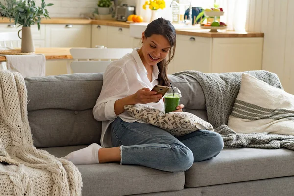 Akıllı Telefon Kullanırken Gülümseyen Mutlu Kadın Evde Yeşil Smoothie Fincan — Stok fotoğraf