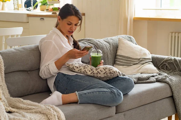 Evde Kanepede Otururken Yeşil Smoothie Içme Akıllı Telefona Bakarak Genç — Stok fotoğraf