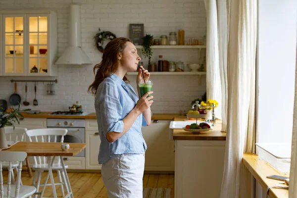 Młoda Kobieta Picie Zielonych Smoothie Stojąc Pobliżu Okna Kuchni — Zdjęcie stockowe