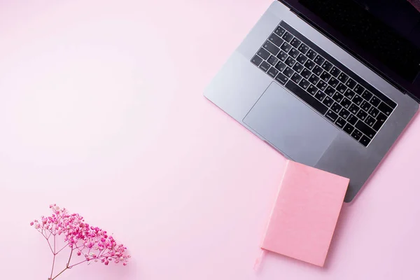 laptop with flowers and notebook on pink background. Top view, blogger workplace concept