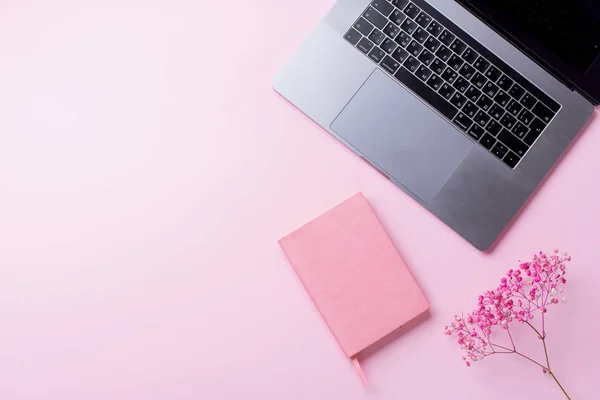 laptop with flowers and notebook on pink background. Top view, blogger workplace concept