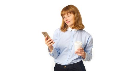 Genç, kızıl saçlı, çilli bir kadın. Elinde kahve fincanı olan akıllı telefon ekranına bakıyor. 