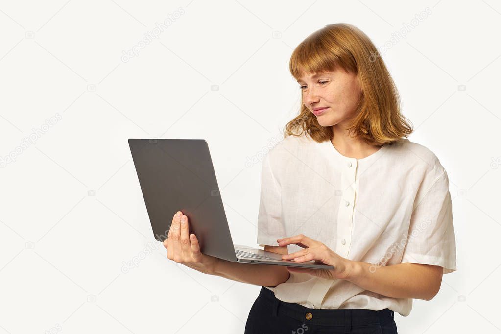 young woman with red hair and freckles holding laptop and looking at screen isolated on white background 