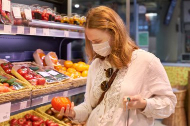 Sarı saçlı, tıbbi koruma maskesi takan bir kadın süpermarkette karantina sırasında domates seçiyor. 