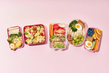 Pembe arka planda çeşitli sağlıklı besleyici yemeklerle birlikte okul beslenme çantalarının üst görüntüsü.