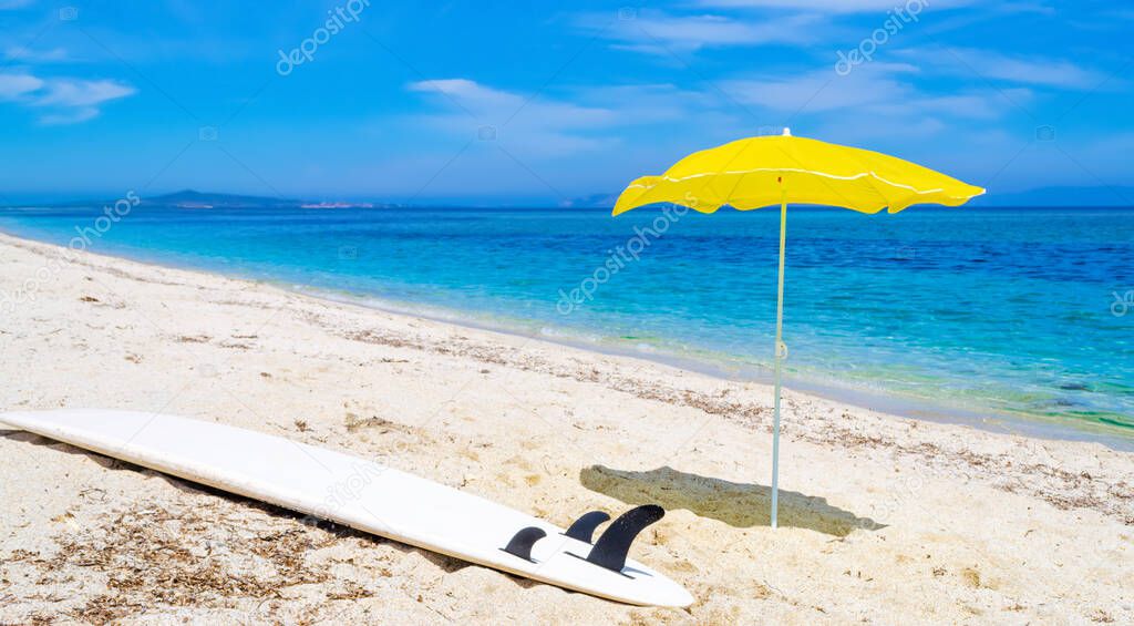 Yellow parasol and a surfboard on a white beach in the summertime