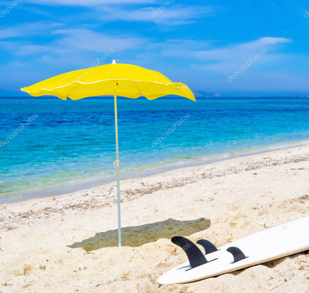 Yellow beach umbrella and a surfboard on a white beach in the summertime