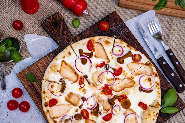 Pizza with chicken, mushrooms, onion and tomatoes, with white sauce. Pizza is on wooden square shaped board with some kitchen objects near