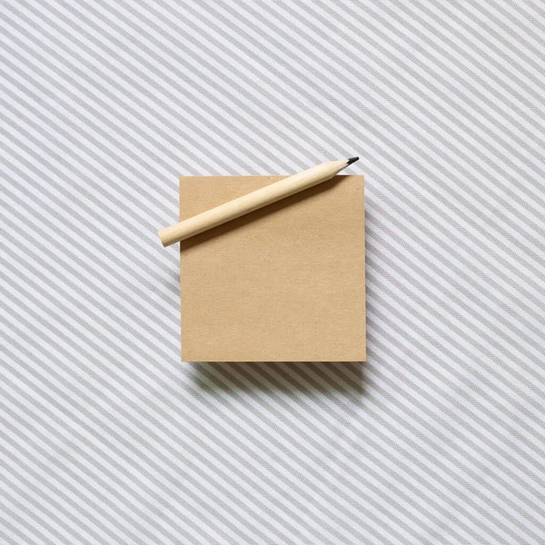 Brown kraft memo pad, empty paper with wooden pencil on gray stripe pattern fabric background. top view, copy space