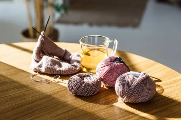 Knitting needles with yarn and knitted textured sweater, hat, scarf are on the table next to a transparent glass cup of hot green tea. Hobbies for pensioners and young mothers