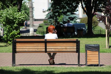 Kızıl saçlı kız şehir parkta bankta oturmuş ve Evet. Kırmızı saç park ve smartphone üzerinde konuşurken bankta oturmuş olan kadın. Kız rahatlatıcı. Kadın bir dinlenme vardır