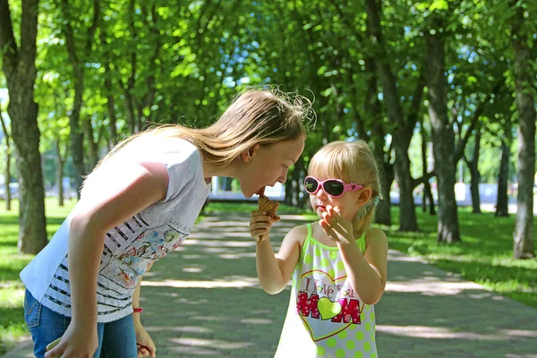 Younger sister giving to eat ice cream to her older sister. Friendship of sisters. Two little sisters walk in summer park and eat ice cream