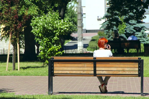 Kızıl saçlı kız şehir parkta bankta oturmuş ve Evet. Kırmızı saç park ve smartphone üzerinde konuşurken bankta oturmuş olan kadın. Kız rahatlatıcı. Kadın bir dinlenme vardır