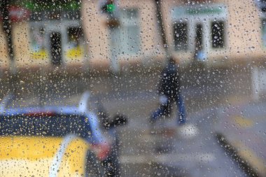 Rain outside window on background of city life. Drops of water dropping on glass during rain. Passers-by pass street in rain. Droplets of water beyond window glass during raining. Rain in city clipart