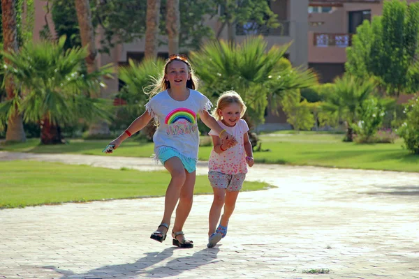 Happy girls run on footpath in tropical resort. Children in palm park. Joyful girls run on path among date palms